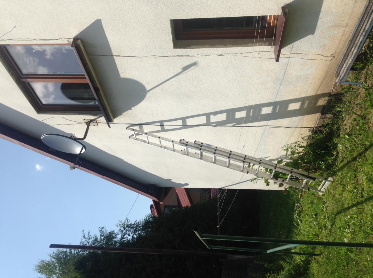 Zamontowana antena satelitarna na elewacji domu, widoczna drabina oparta o ścianę i cień anteny.