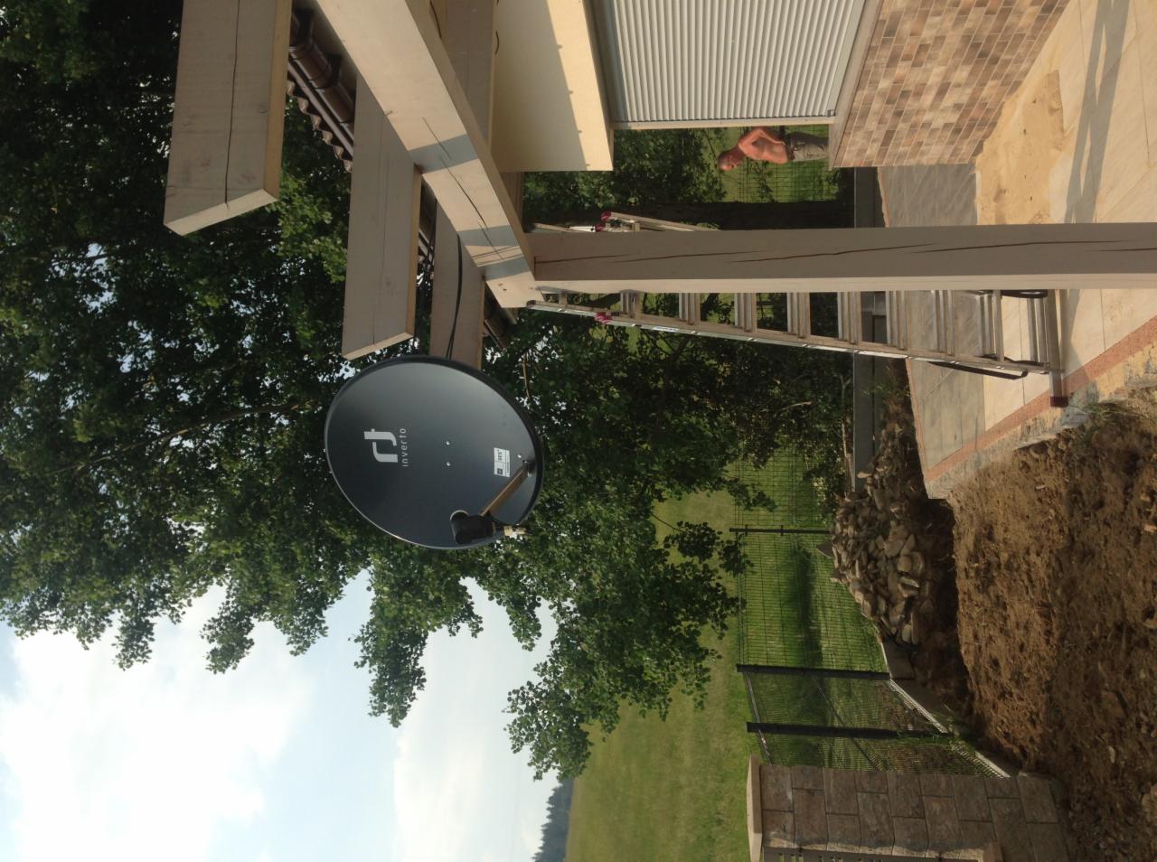 Czarna antena satelitarna marki Inverto zamontowana na drewnianej konstrukcji, widok z dołu na tle zieleni i nieba, drabina oparta o konstrukcję, mur z cegły w tle.