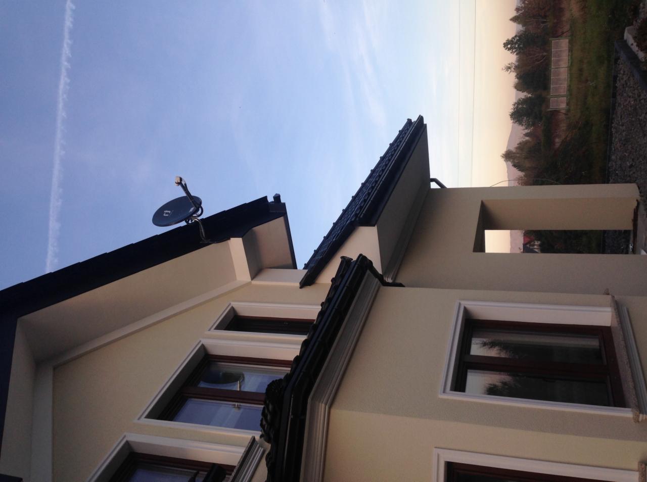 Antena satelitarna zamontowana na dachu domu jednorodzinnego, widok z dołu na tle błękitnego nieba i zachodzącego słońca.