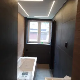 Nowoczesna łazienka z szarymi płytkami, oświetleniem LED w suficie i białą wanną oraz umywalką. Widoczne okno z widokiem na budynki.