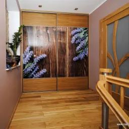 Korytarz z drewnianą podłogą i przesuwną szafą z frontami imitującymi drewno i motywem lawendy, widoczna drewniana balustrada schodów i drzwi z elementami szkła.