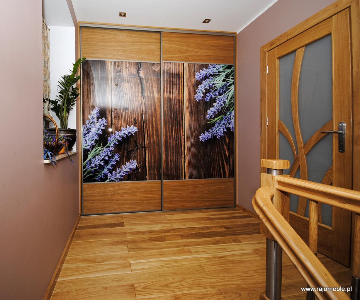 Korytarz z drewnianą podłogą i przesuwną szafą z frontami imitującymi drewno i motywem lawendy, widoczna drewniana balustrada schodów i drzwi z elementami szkła.