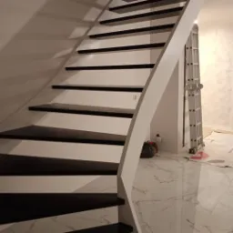 Schody drewniane z czarnymi stopniami i białą balustradą, widoczne narzędzia i drabina w tle, marmurowa podłoga.