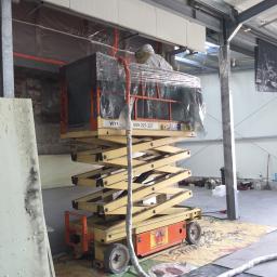 Usługi Remontowo Budowlane IZOBUD Bodnik Krzysztof - Pracownik w kombinezonie ochronnym na podnośniku nożycowym, zabezpieczający folią fragment konstrukcji hali przed aplikacją izolacji natryskowej; widoczna płyta izolacyjna i osłonięte elementy hali.