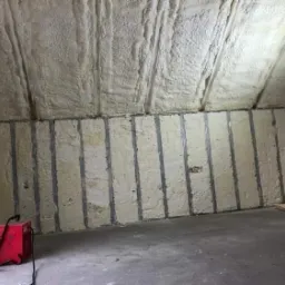 Poddasze z widoczną izolacją pianką natryskową na skośnym suficie i panelami izolacyjnymi z metalowymi profilami pionowymi na ścianie, czerwony agregat natryskowy stoi na betonowej podłodze.