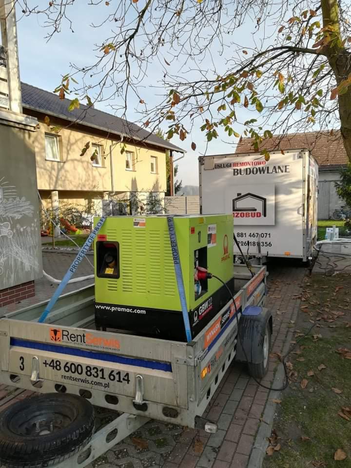 Agregat prądotwórczy marki Pramac GBW22 na przyczepie Rent Serwis, zaparkowany obok przyczepy z reklamą usług remontowo-budowlanych IZOBUD na tle domu jednorodzinnego.