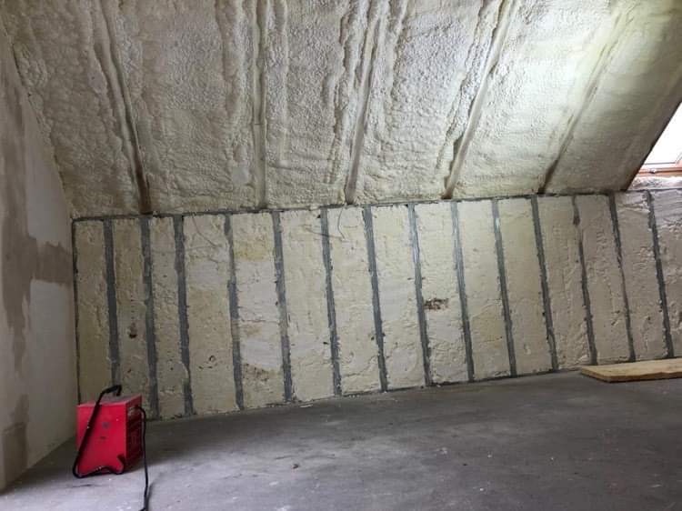 Poddasze z widoczną izolacją pianką natryskową na skośnym suficie i panelami izolacyjnymi z metalowymi profilami pionowymi na ścianie, czerwony agregat natryskowy stoi na betonowej podłodze.