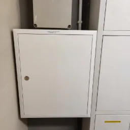 Biała skrzynka elektryczna z zamkiem na ścianie, widoczne przewody i rury, obok szafka z napisem 'Ochronnik przepięciowy'.