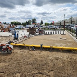 Maskin Roboty Ziemne - Dwóch pracowników układa kostkę brukową na przygotowanym podłożu z użyciem żółtej listwy wyrównującej, w tle widoczne zaparkowane samochody i stacja benzynowa.