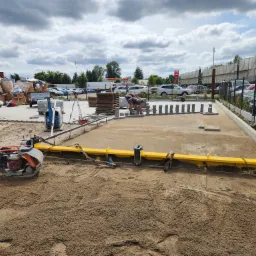 Dwóch pracowników układa kostkę brukową na przygotowanym podłożu z użyciem żółtej listwy wyrównującej, w tle widoczne zaparkowane samochody i stacja benzynowa.