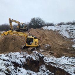 Maskin Roboty Ziemne - Dwie żółte koparki Komatsu pracują na zaśnieżonym terenie, usypując górę ziemi. Jedna koparka stoi na szczycie, druga na pierwszym planie. Pochmurne niebo.