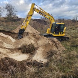 Maskin Roboty Ziemne - Żółta koparka Komatsu w trakcie prac ziemnych, wykop pod fundamenty na nierównym terenie z suchą trawą i drzewami w tle, pochmurne niebo.