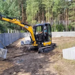 Żółta koparka JCB montuje panel ogrodzenia betonowego w Wałczu. Gotowe segmenty ogrodzenia w tle, drzewa i trawa. Operator w kabinie daje znak ręką.