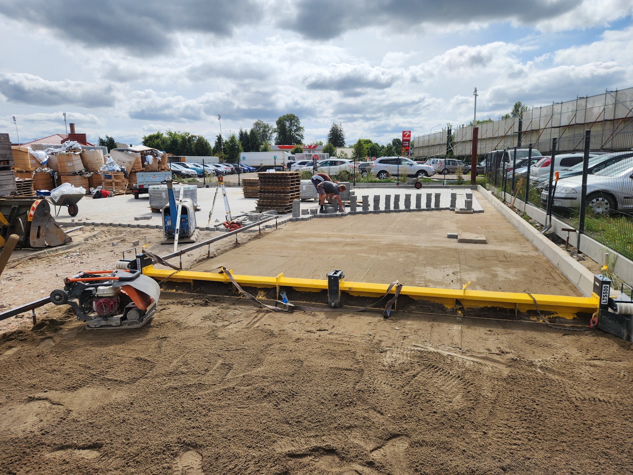 Dwóch pracowników układa kostkę brukową na przygotowanym podłożu z użyciem żółtej listwy wyrównującej, w tle widoczne zaparkowane samochody i stacja benzynowa.