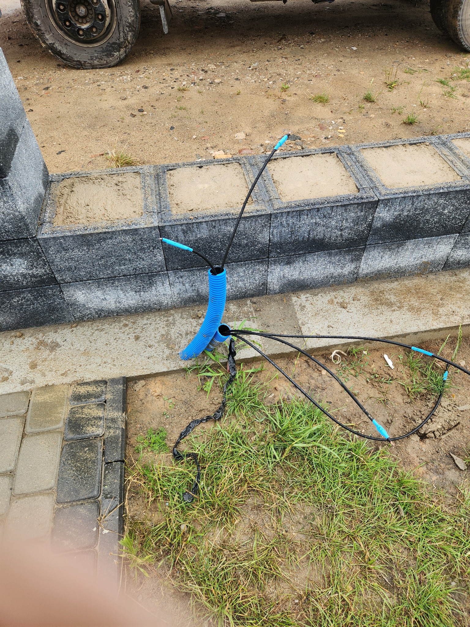 Fragment budowy: niebieska rura karbowana wystaje z ziemi obok betonowego murku i kostki brukowej, z niej wychodzą czarne kable z niebieskimi końcówkami.