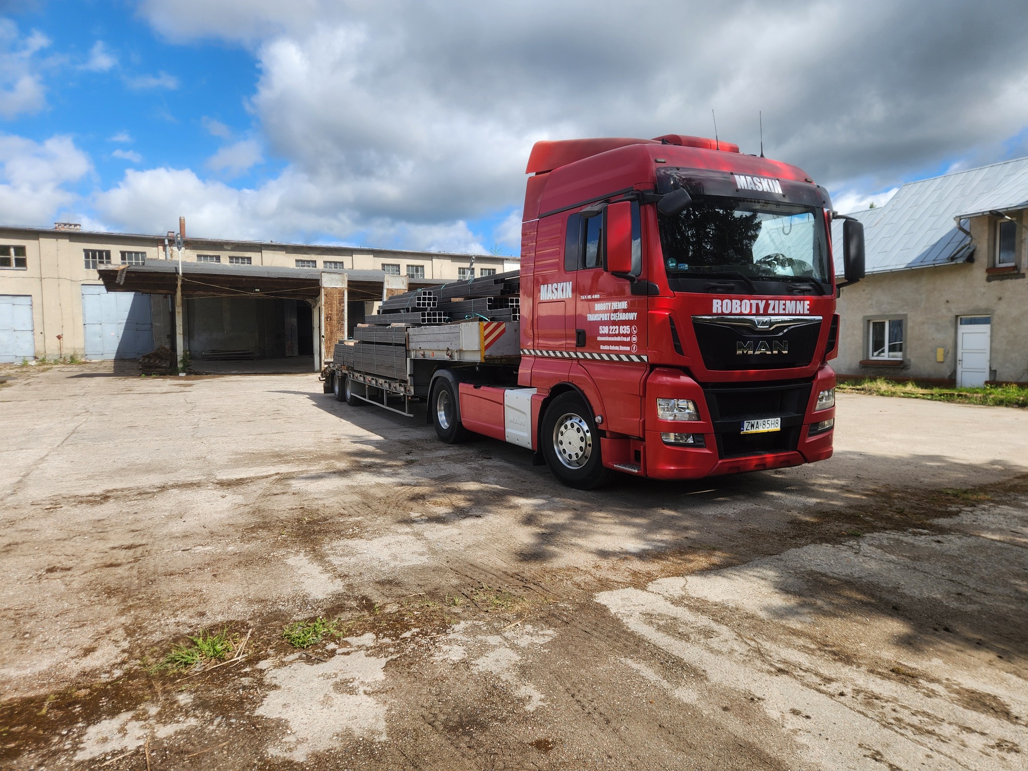 Czerwona ciężarówka MAN z napisem 'ROBOTY ZIEMNE' i nazwą firmy, załadowana stalowymi profilami, stoi na placu przed budynkiem magazynowym.
