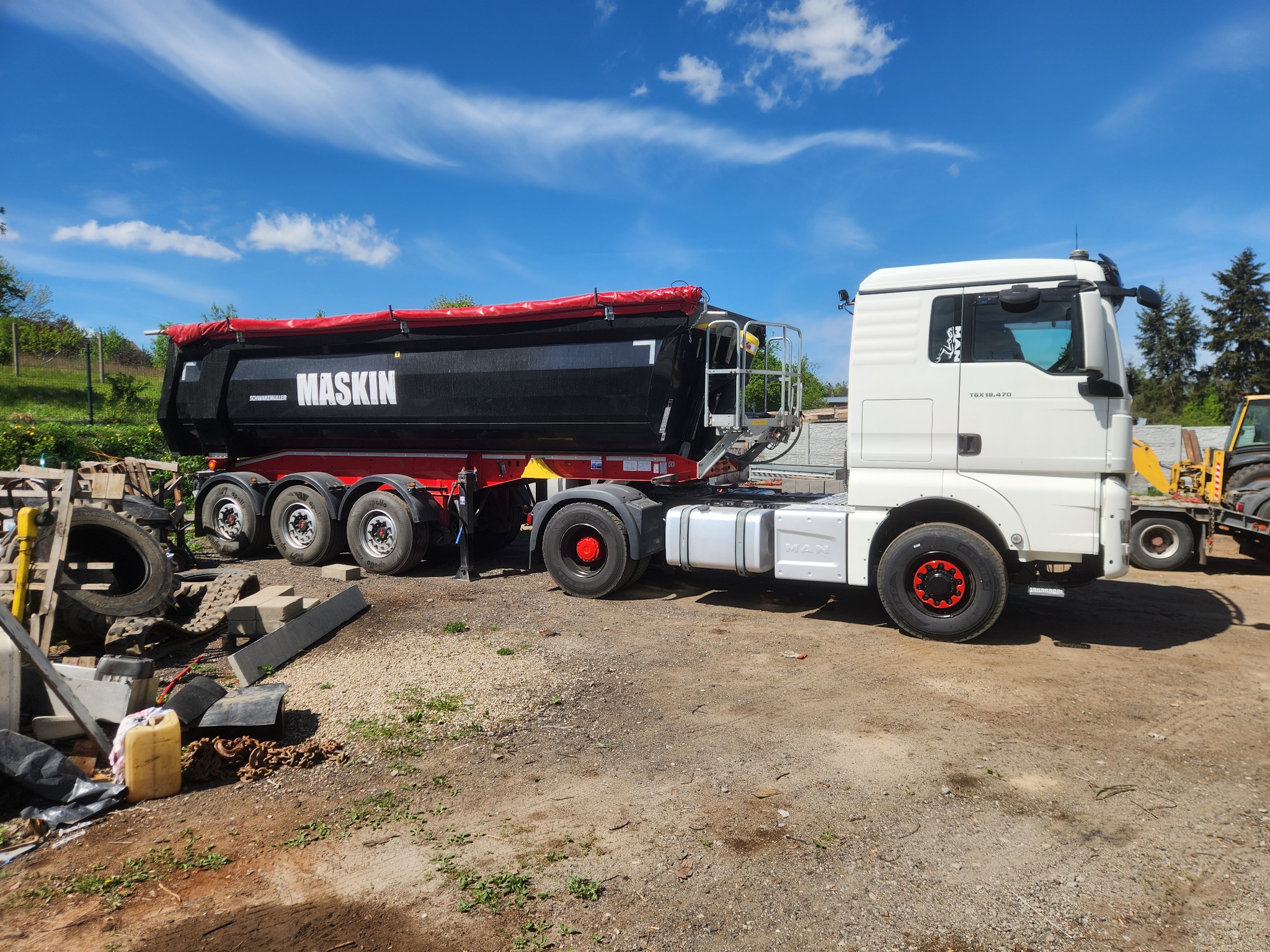 Ciężarówka MAN z naczepą wywrotką Maskin na placu budowy, w tle błękitne niebo i maszyny budowlane. Widoczne opony i elementy konstrukcyjne naczepy.