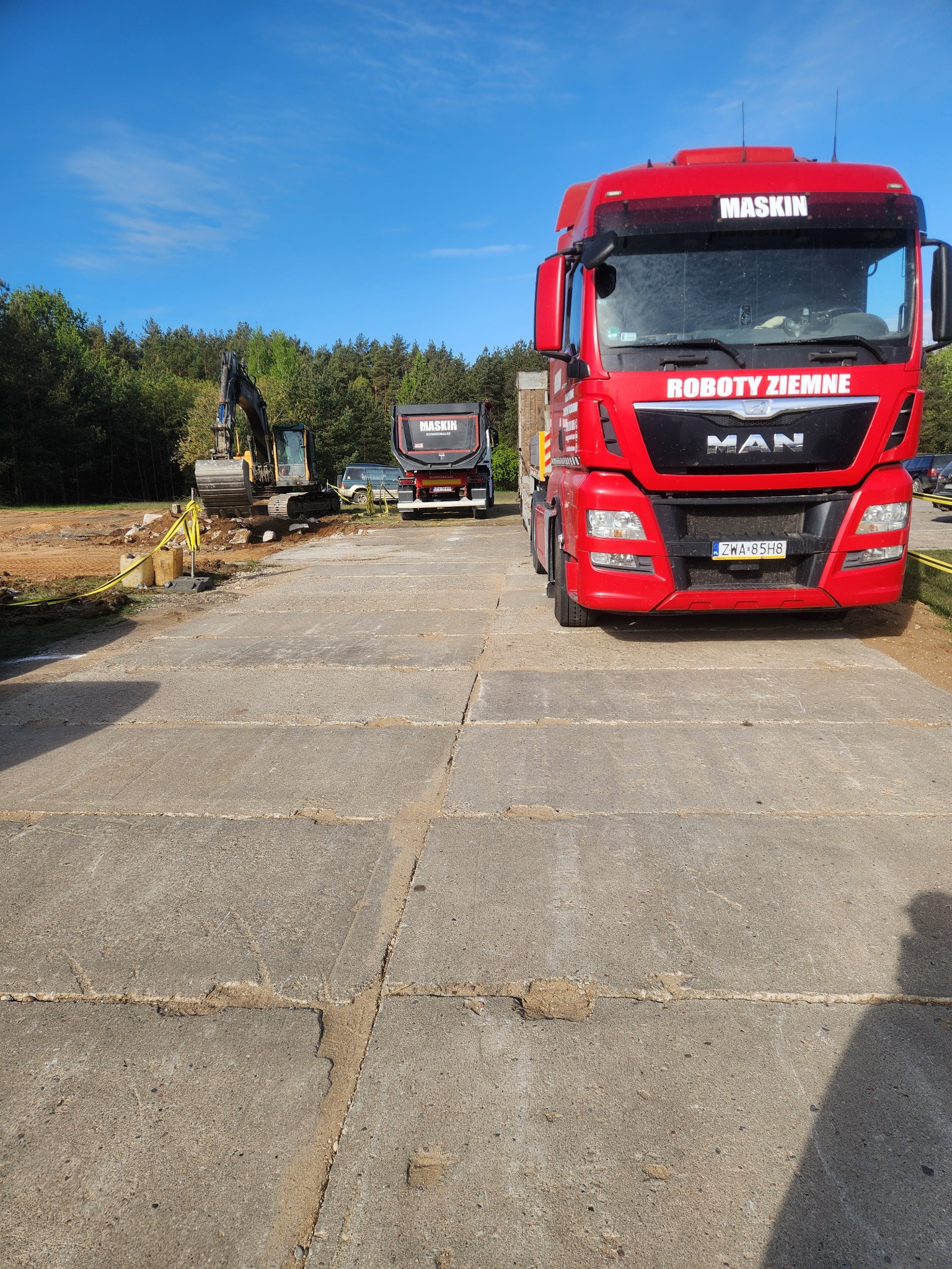 Czerwona ciężarówka MAN z napisem 'ROBOTY ZIEMNE' na tle wykopu i koparki, na betonowej nawierzchni. Widoczne maszyny firmy Maskin.