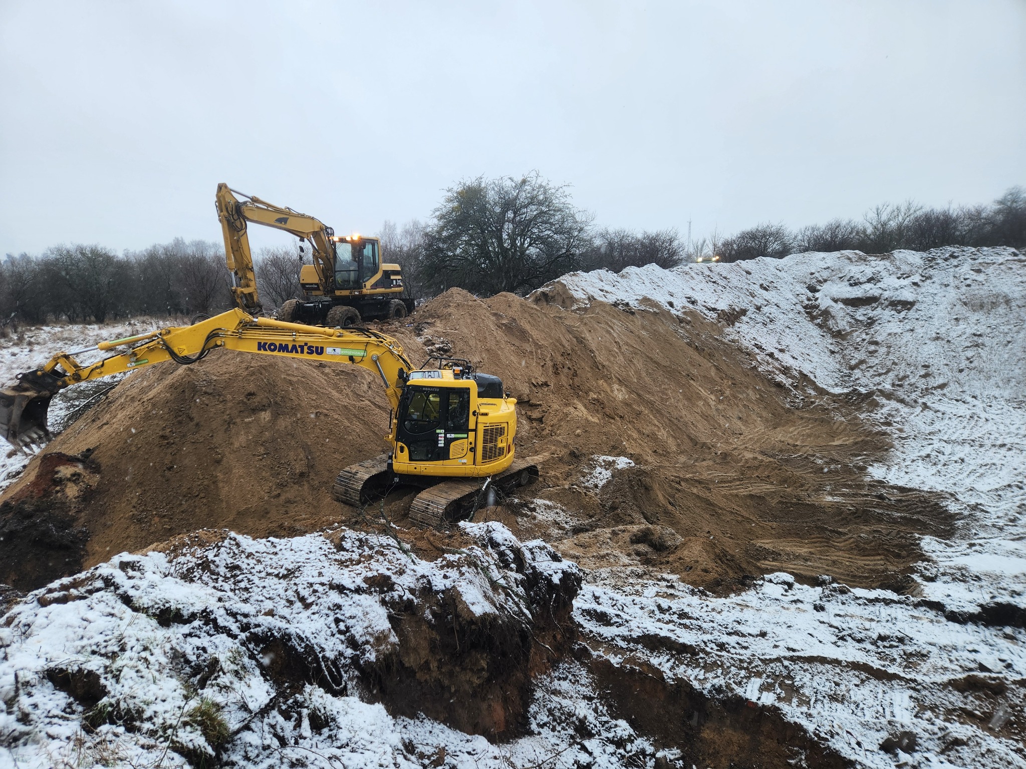 Dwie żółte koparki Komatsu pracują na zaśnieżonym terenie, usypując górę ziemi. Jedna koparka stoi na szczycie, druga na pierwszym planie. Pochmurne niebo.