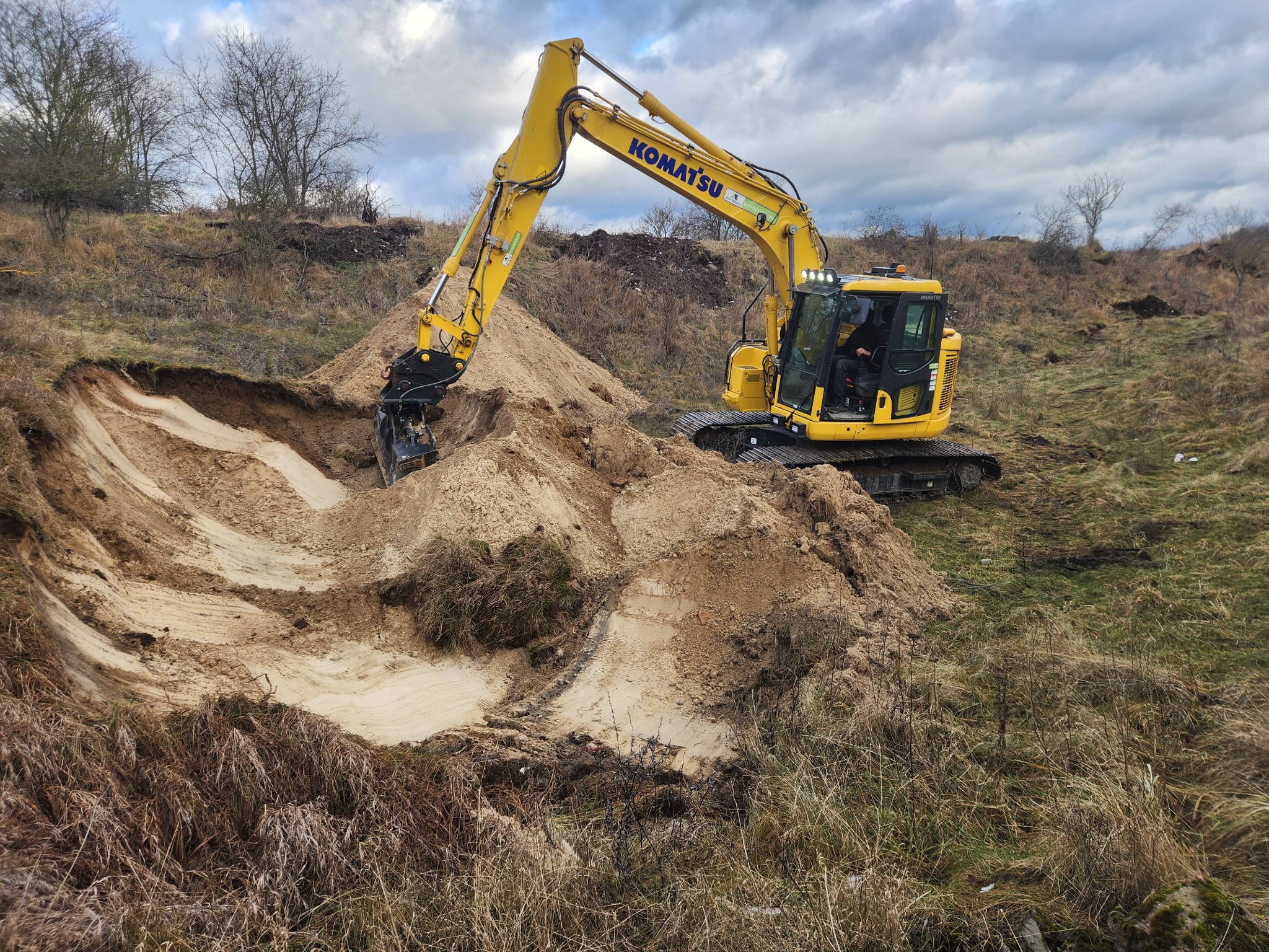 Żółta koparka Komatsu w trakcie prac ziemnych, wykop pod fundamenty na nierównym terenie z suchą trawą i drzewami w tle, pochmurne niebo.