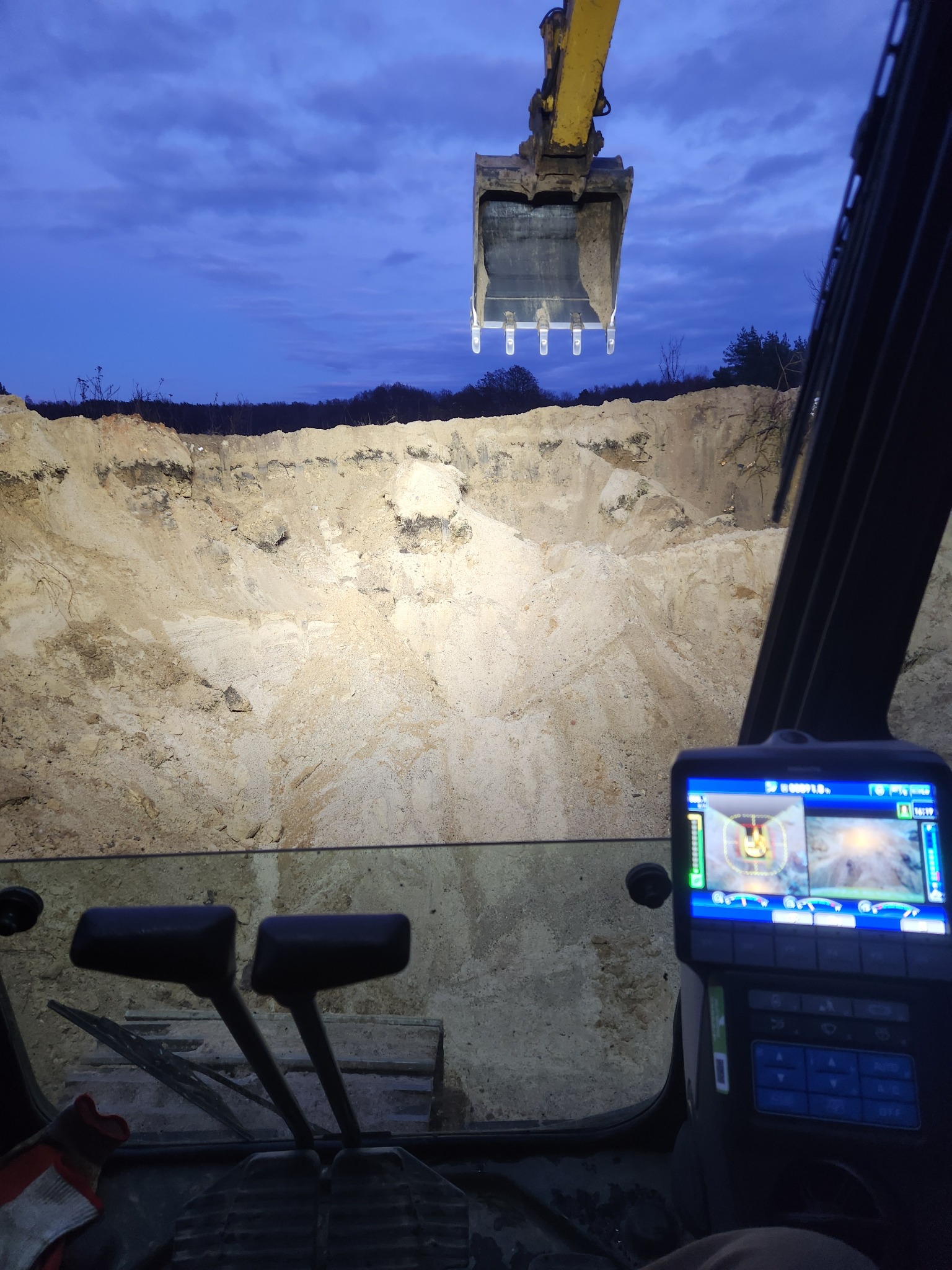 Widok z kabiny koparki na wykop oświetlony reflektorami, z łyżką koparki w górnej części kadru na tle wieczornego nieba. Widoczny panel sterowania maszyny.
