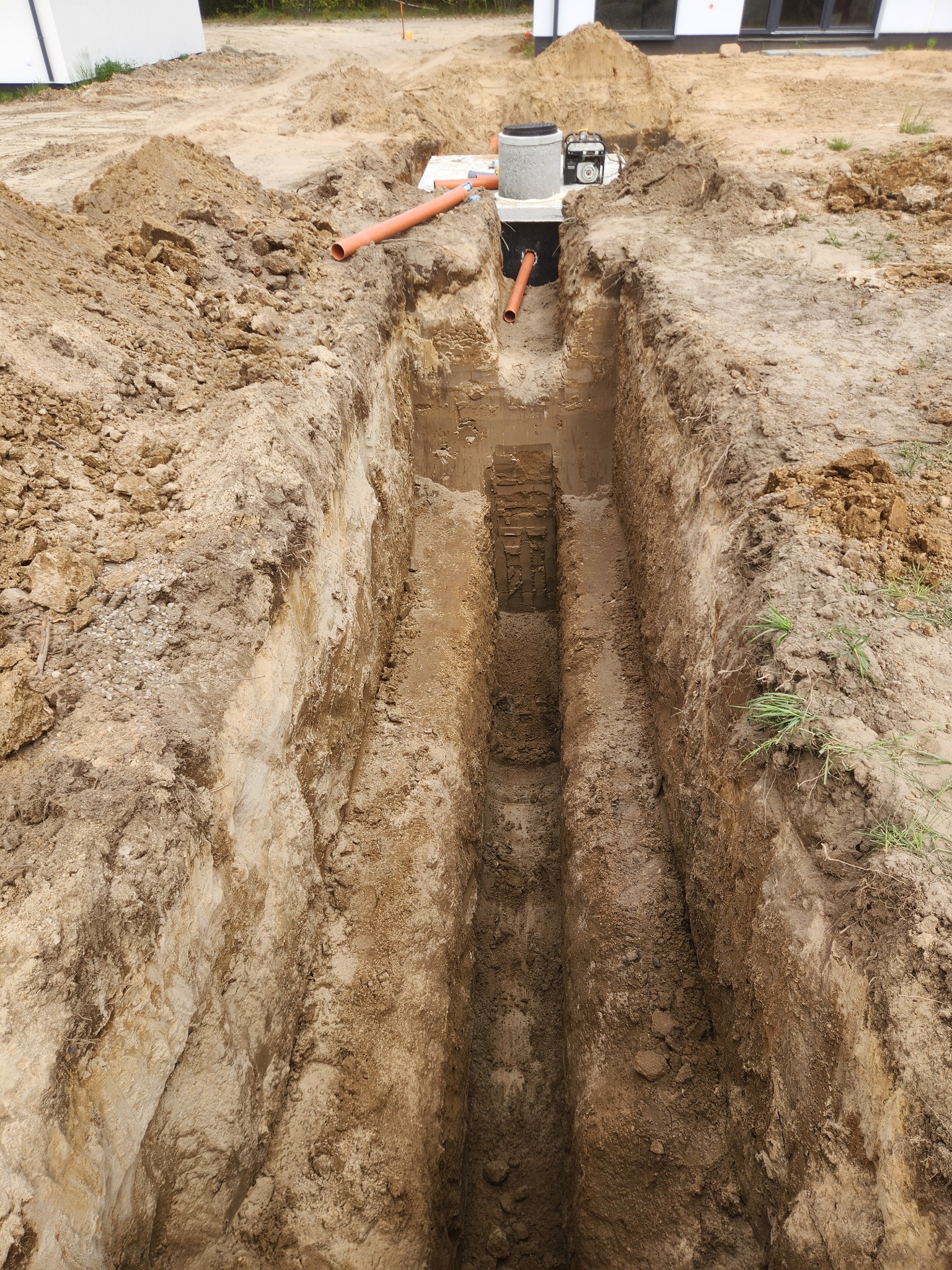 Wykopany głęboki rów pod instalację kanalizacyjną z widocznym betonowym zbiornikiem i rurami, ziemia usypana wokół. Etap prac ziemnych.