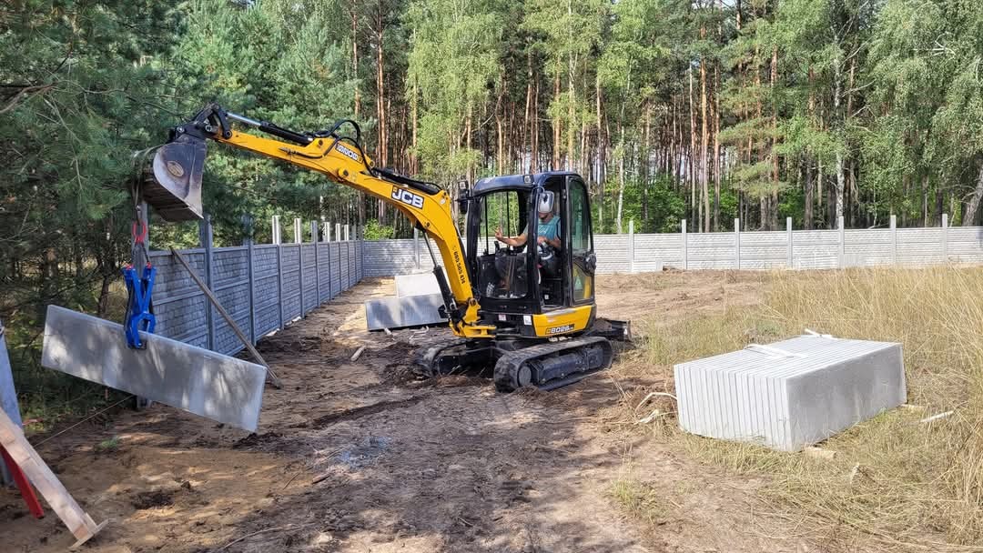 Żółta koparka JCB montuje panel ogrodzenia betonowego w Wałczu. Gotowe segmenty ogrodzenia w tle, drzewa i trawa. Operator w kabinie daje znak ręką.