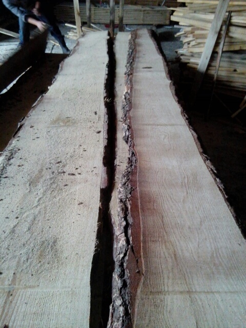 Dwie szerokie deski tarcicy z korą po bokach, ułożone równolegle, widoczne ślady obróbki, w tle stos desek i osoba przenosząca drewno.