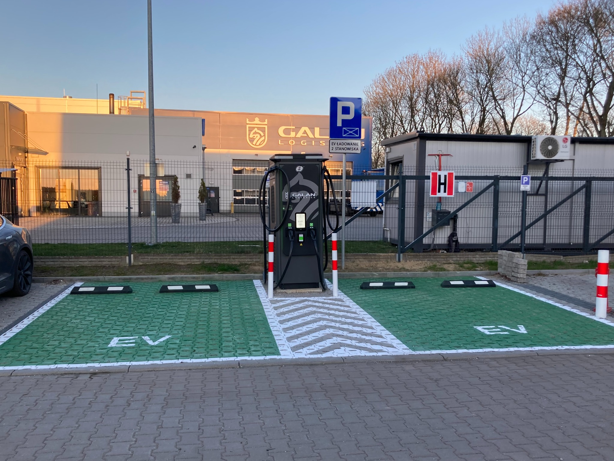 Stacja ładowania EV z dwoma stanowiskami, zielone miejsca parkingowe z napisem 'EV', budynek w tle oświetlony słońcem, znak 'P' i 'H' na terenie.