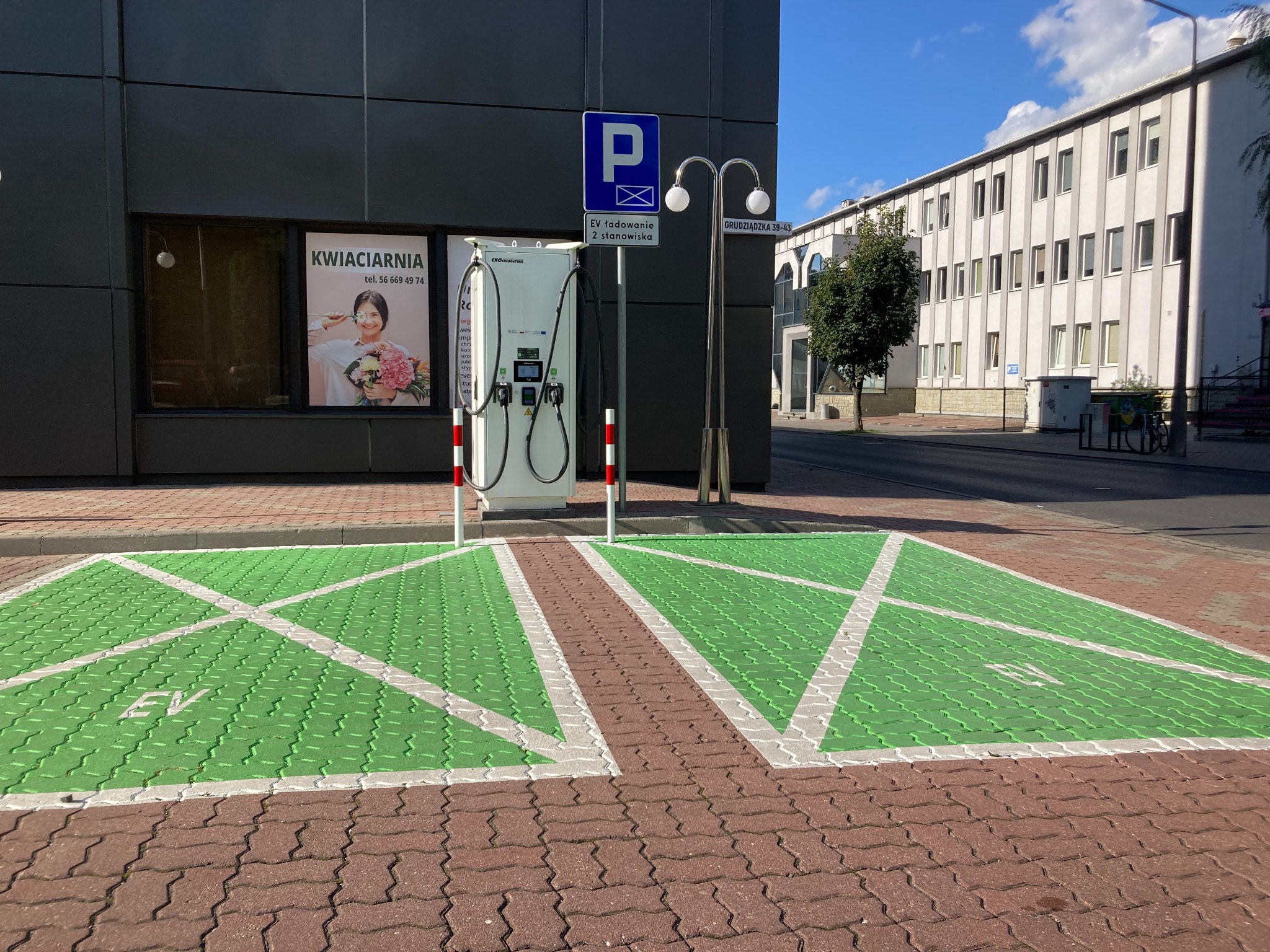 Stacja ładowania EV z dwoma stanowiskami, oznaczona zielonymi i białymi pasami na kostce brukowej. W tle budynek z reklamą kwiaciarni i tabliczka z adresem.