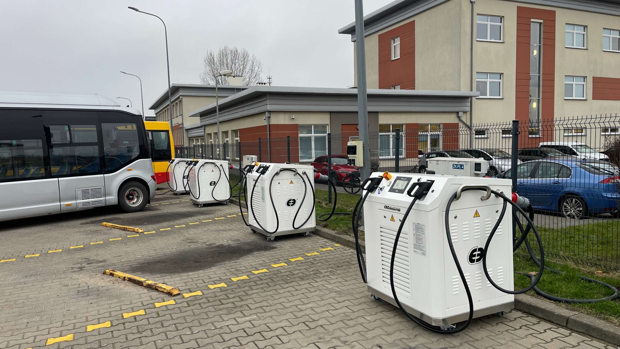 Mobilne stacje ładowania autobusów elektrycznych ustawione w rzędzie na zewnątrz budynku. Widoczne kable i złącza. Szara kostka brukowa i żółte oznaczenia.