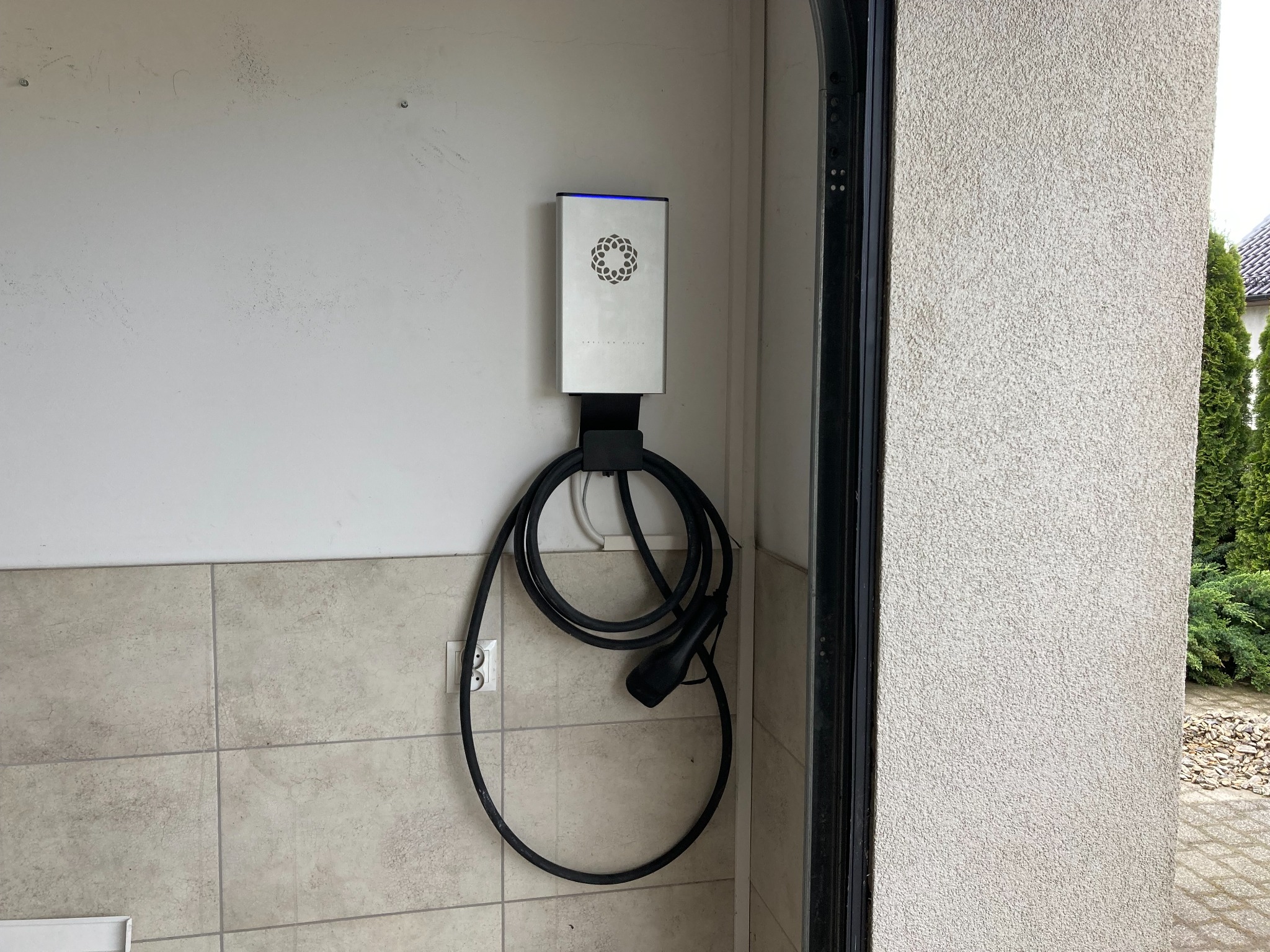 Zamontowana na ścianie ładowarka EV Wallbox z kablem, w garażu wykończonym płytkami. Widoczny fragment bramy garażowej i zewnętrzny ogród.