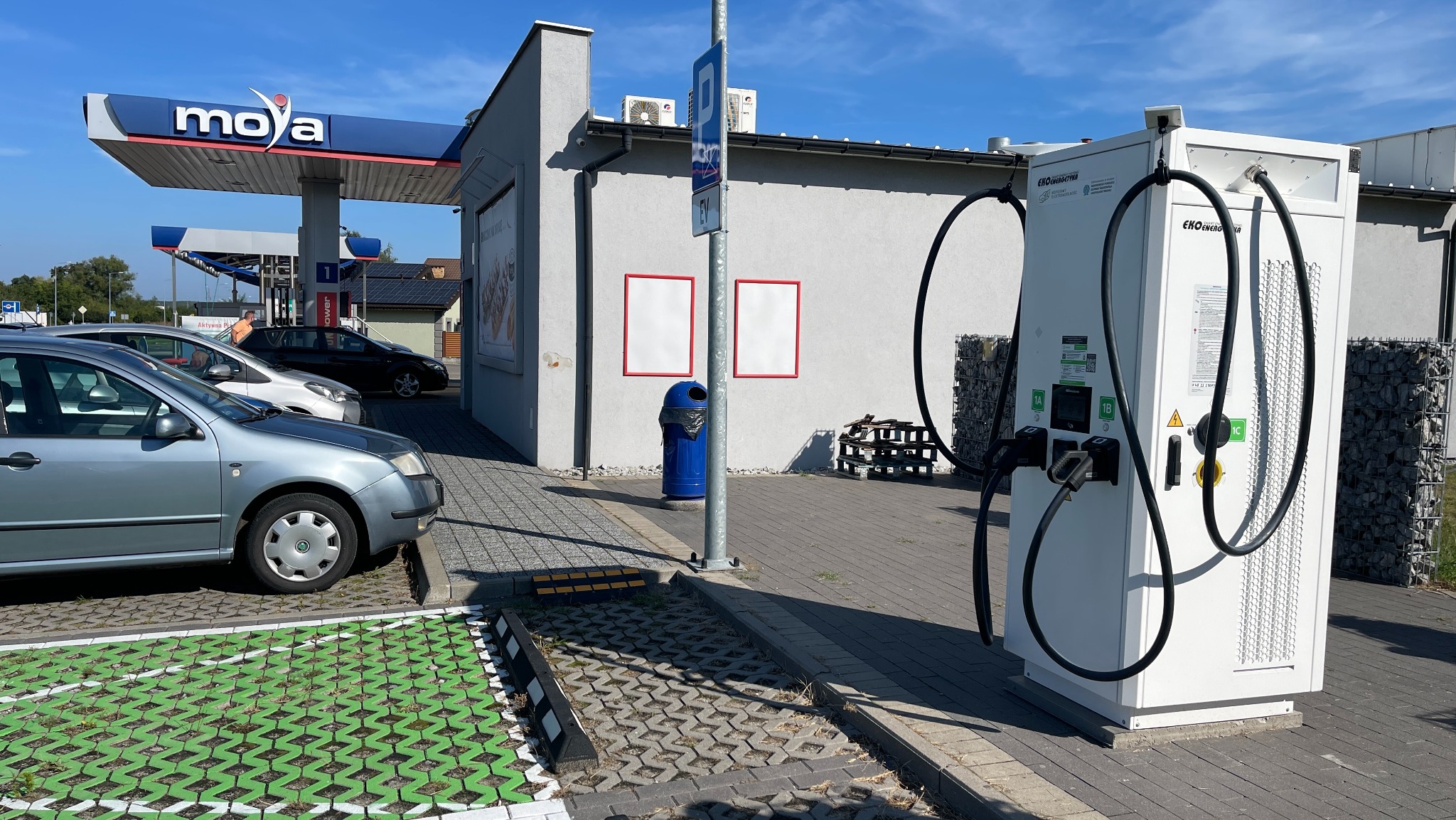 Stacja ładowania samochodów elektrycznych Ekoen Energetyka na stacji paliw Moya, z zaparkowanym szarym samochodem osobowym i zielonymi miejscami parkingowymi.