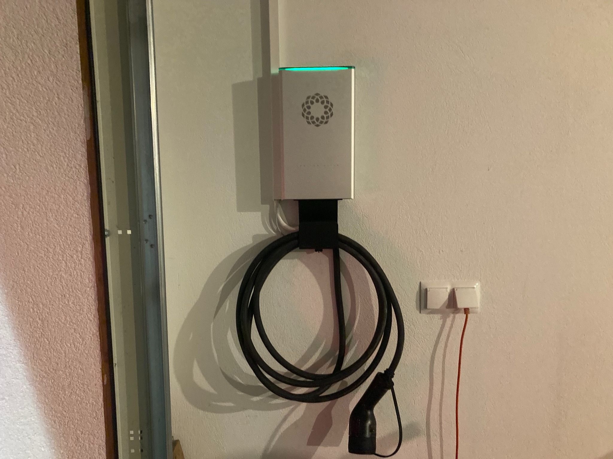 Nowoczesna stacja ładowania pojazdów elektrycznych zamontowana na ścianie w garażu, z widocznym kablem i gniazdkiem. Górna część podświetlona na zielono.