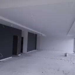 Wnętrze w trakcie wykańczania: ściany w kolorze grafitowym i białe, gładkie sufity z wystającymi przewodami, podłoga zabezpieczona folią malarską, widoczny pojemnik z farbą w rogu pomieszczenia.