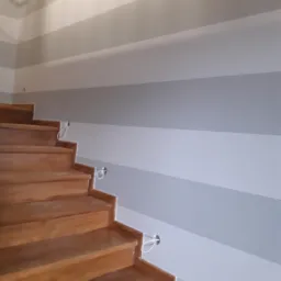 Drewniane schody z pomalowaną na szaro-białe pasy ścianą w tle, widoczne przewody elektryczne przy ścianie.