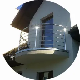 Balkon z zaokrągloną, nowoczesną balustradą ze stali nierdzewnej, z widocznymi refleksami słońca na metalowych elementach, na tle elewacji domu.