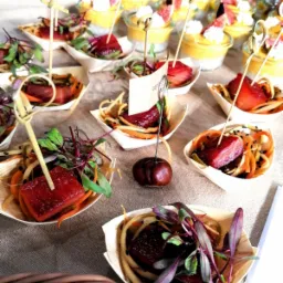 Elegancko podane przekąski cateringowe: kostki mięsa na warzywnych spaghetti w łódeczkach z jasnego drewna, udekorowane zieleniną i szpikulcem, w tle desery w szklankach z owocami.