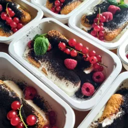 Porcje tiramisu w białych, plastikowych pojemnikach na wynos, udekorowane malinami, czerwoną porzeczką i listkiem mięty.