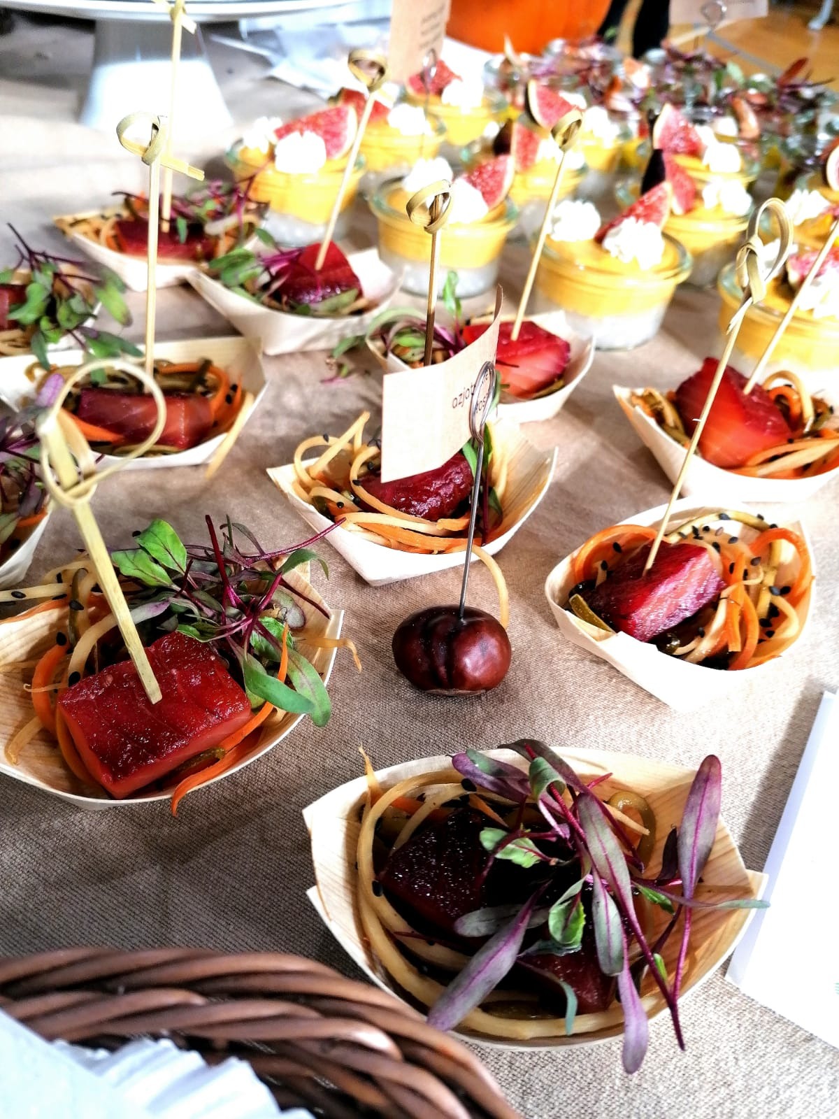 Elegancko podane przekąski cateringowe: kostki mięsa na warzywnych spaghetti w łódeczkach z jasnego drewna, udekorowane zieleniną i szpikulcem, w tle desery w szklankach z owocami.