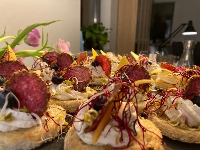 Dekoracyjne kanapeczki z salami, serem, borówkami i kiełkami na jasnym pieczywie, ułożone na ciemnej powierzchni, z tulipanami i lampą w tle.