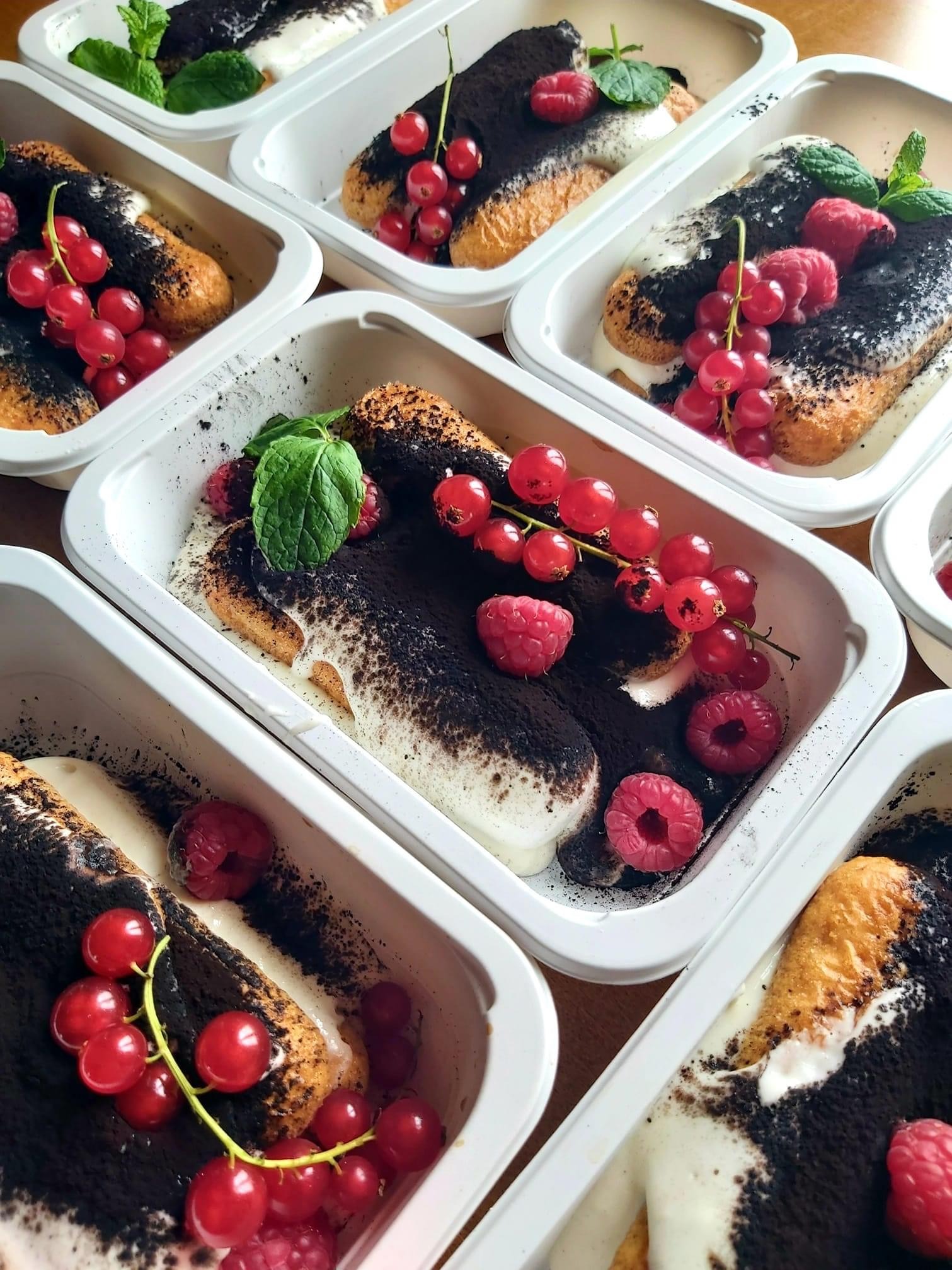 Porcje tiramisu w białych, plastikowych pojemnikach na wynos, udekorowane malinami, czerwoną porzeczką i listkiem mięty.