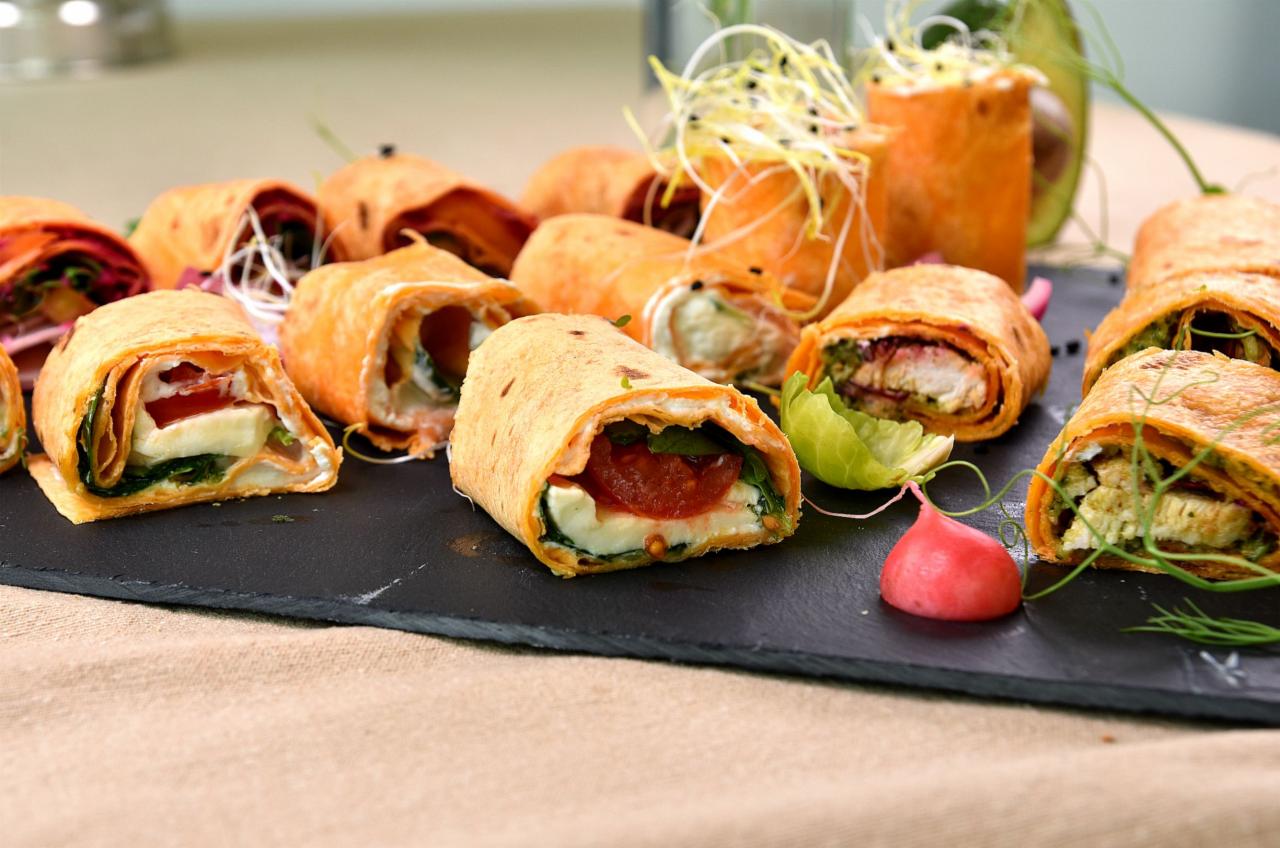 Tortilla wrap przekrojona na pół, ukazująca wnętrze z mozzarellą, pomidorem i szpinakiem, ułożona na czarnym kamiennym talerzu obok innych wrapów z różnymi nadzieniami i kiełkami.