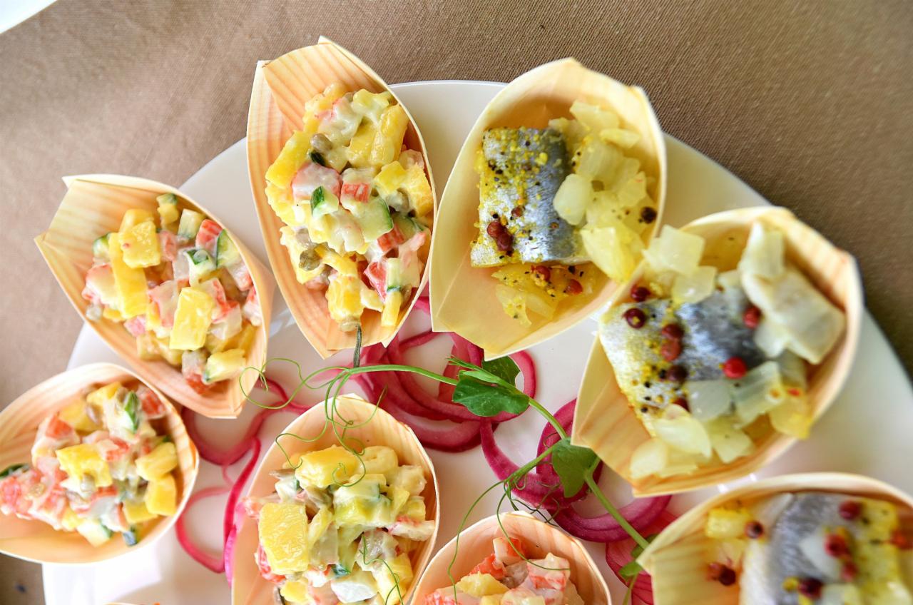 Dekoracyjne łódeczki z drewna wypełnione sałatką jarzynową i śledziem w oleju na białym talerzu, ozdobione piórami czerwonej cebuli i pędami groszku.