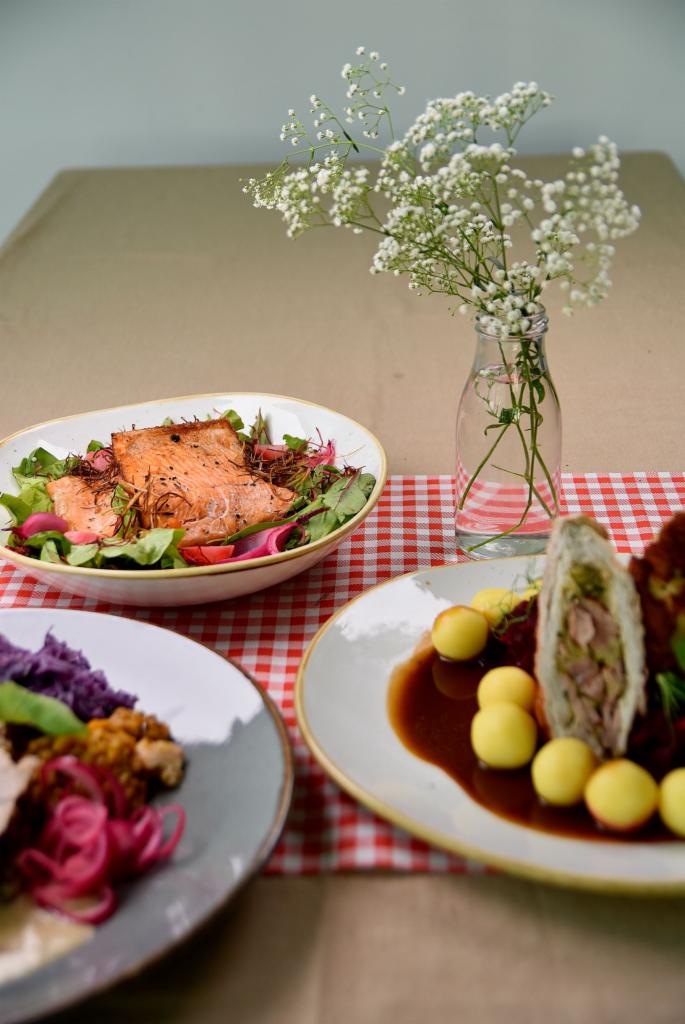 Trzy talerze z różnorodnymi daniami na stole pokrytym czerwoną kratą, w tle wazonik z drobnymi białymi kwiatami i beżowy obrus.