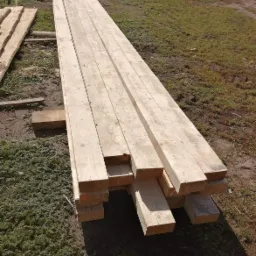 Stos jasnej, surowej kantówki ułożonej na podkładach z drewna na trawiastym podłożu, widoczne ślady cięcia i naturalne niedoskonałości drewna.