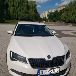Biała Skoda Superb na brukowanym placu z widokiem na drzewa i budynki w tle, z zawieszką zapachową w kształcie choinki na lusterku wstecznym.