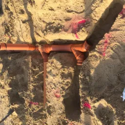 Pomarańczowe rury kanalizacyjne ułożone w wykopie w piaszczystym gruncie, widoczne ślady czerwonej farby geodezyjnej i butelka z klejem do rur.