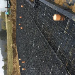 Siatka zbrojeniowa na czarnej folii, zabezpieczona plastikowymi opaskami, z wystającymi rurkami PVC w kolorze pomarańczowym, przygotowana do zalania betonem, widok z boku na tle krajobrazu.