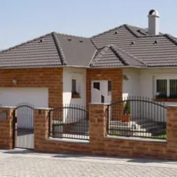 Jednopiętrowy dom z dwuspadowym dachem z brązowej dachówki, elewacją z cegły klinkierowej i białymi elementami, czarny metalowy płot z ceglanymi słupkami, brama wjazdowa, widok z zewnątrz...